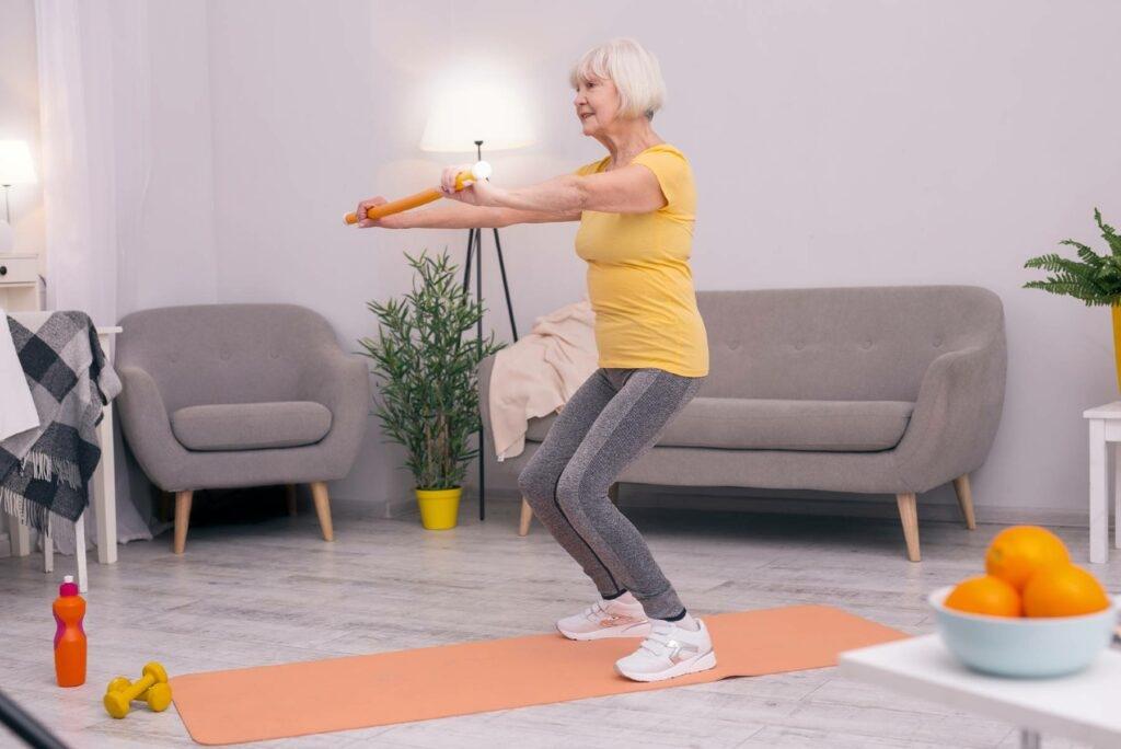 A senior woman exercises in her living room with a balance bar and a yoga mat