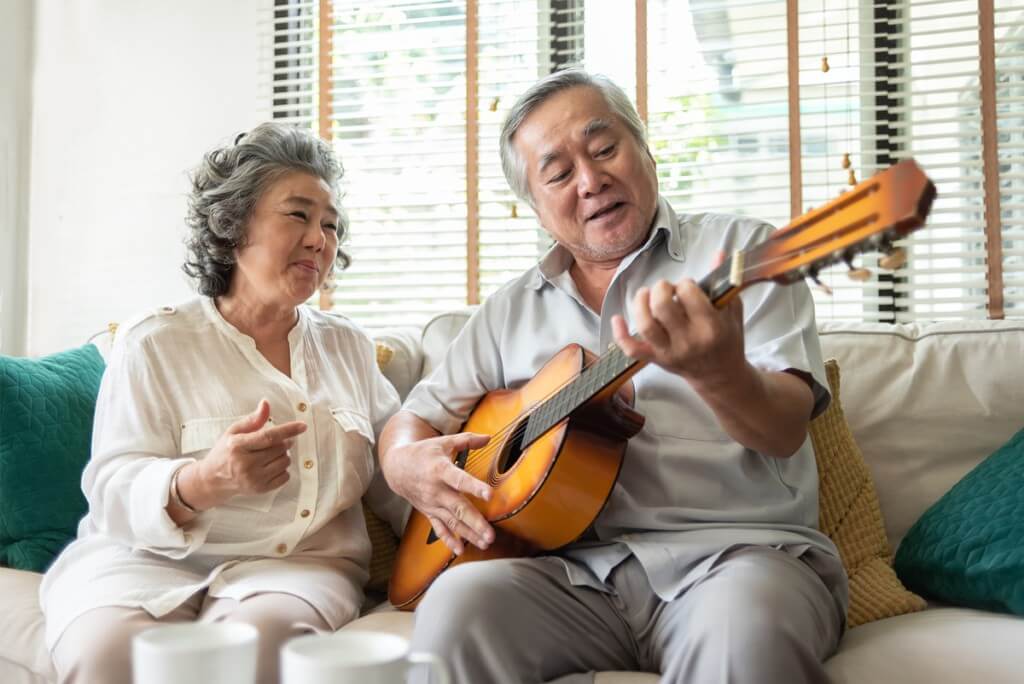 A senior man and a senior woman sit on the couch together the woman sings and the man accompanies her on a guitar