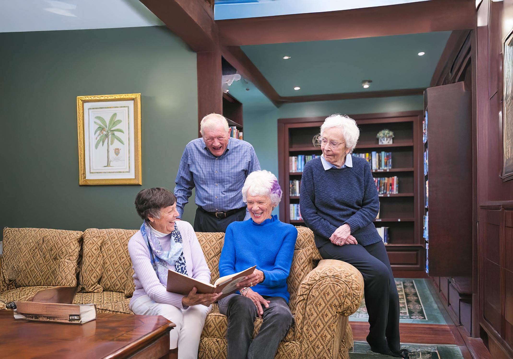 Five Meadow Ridge residents gather in the community library, smiling and reading together on a sofa surrounded by bookshelves, capturing a warm moment of connection and shared learning.