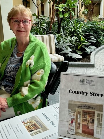 A Meadow Ridge resident wearing a green cardigan sits at a table indoors beside a sign for the Meadow Ridge Country Store, with plants and seating visible in the background, highlighting resident involvement and support of the community fundraiser.