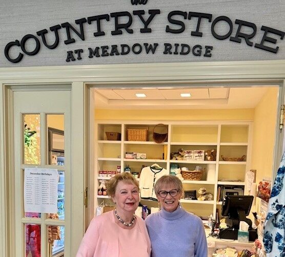Two smiling women stand in the doorway of The Country Store at Meadow Ridge, with shelves of gifts and everyday items visible behind them inside the warmly lit shop.