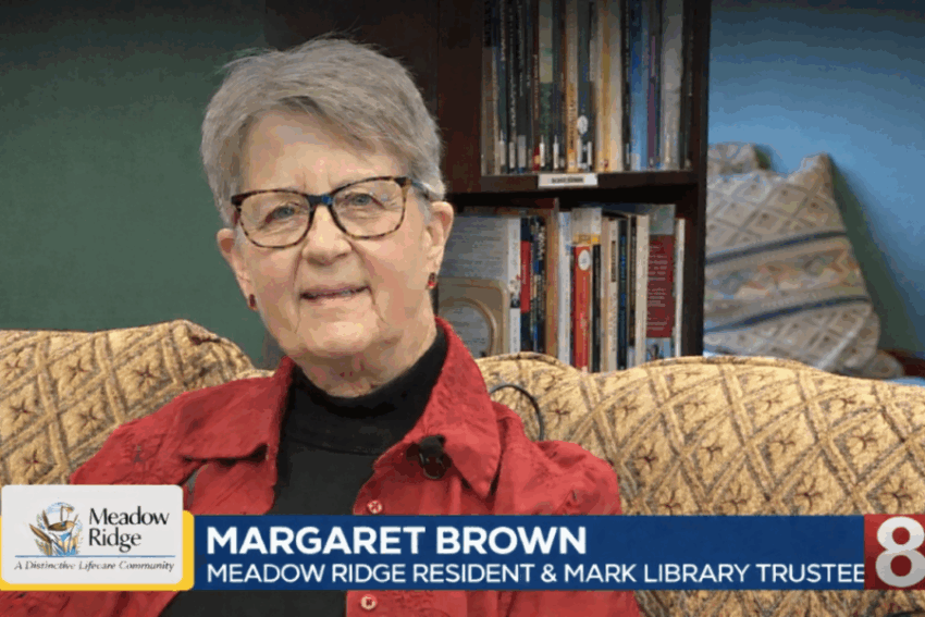 Margaret Brown, a Meadow Ridge resident and Mark Twain Library trustee, sits on a patterned sofa wearing a red jacket and black turtleneck, smiling gently toward the camera, with bookshelves in the background and an on-screen lower-third identifying her name and role alongside the Meadow Ridge and Channel 8 logos.
