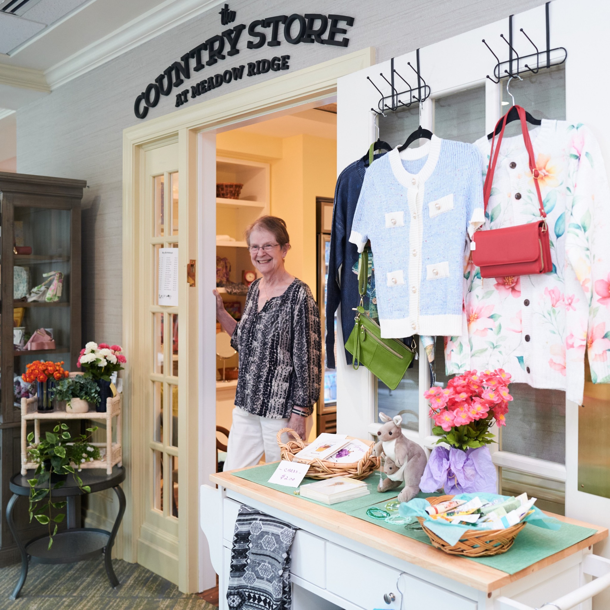 Laurie S., a Meadow Ridge resident volunteer, smiles while standing in the doorway of the Country Store, surrounded by colorful clothing, handbags, plants, and gift items that help keep the store welcoming and well stocked.