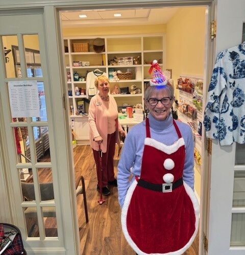 A Meadow Ridge resident volunteer wearing a festive red holiday apron and hat smiles while standing in the doorway of the Country Store, with another resident visible inside browsing shelves filled with gifts and merchandise.