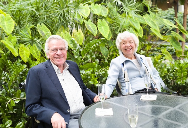 Older man and woman smile while seated at an outdoor patio table surrounded by lush greenery, with champagne flutes in front of them.