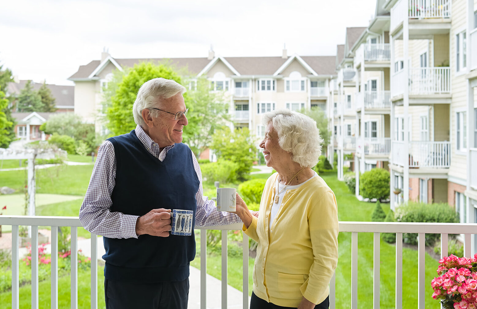 Older couple standing on a balcony at a senior living community, smiling and talking while holding coffee mugs. Apartment-style buildings and green landscaping are visible in the background.