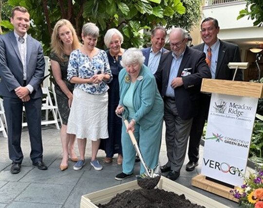 Susan Auslander, a Meadow Ridge resident, ceremonially breaks ground with a shovel in front of a raised bed filled with soil while community leaders and guests stand behind her smiling during an indoor sustainability event. A podium beside the group displays Meadow Ridge, Connecticut Green Bank and Verogy signage.