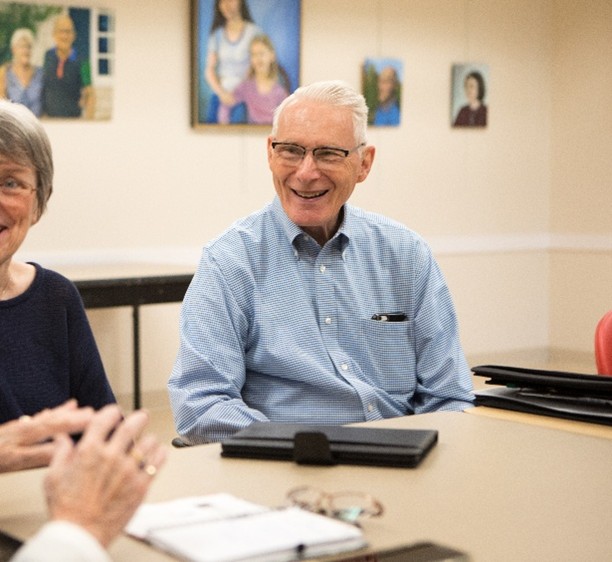 Older man in a light blue button-down shirt smiles during a meeting around a conference table, with other residents partially visible and framed portraits hanging on the wall behind him.