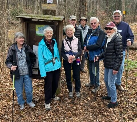 A group of older adults stand on a wooded trail at a preserve, smiling and holding walking sticks beside a trailhead sign, surrounded by leaf-covered ground and tall trees.