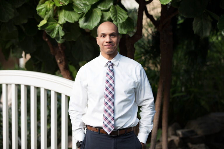 A professionally dressed man stands outdoors near a white footbridge, wearing a white dress shirt, patterned tie, and dark slacks, with greenery and trees in the background.