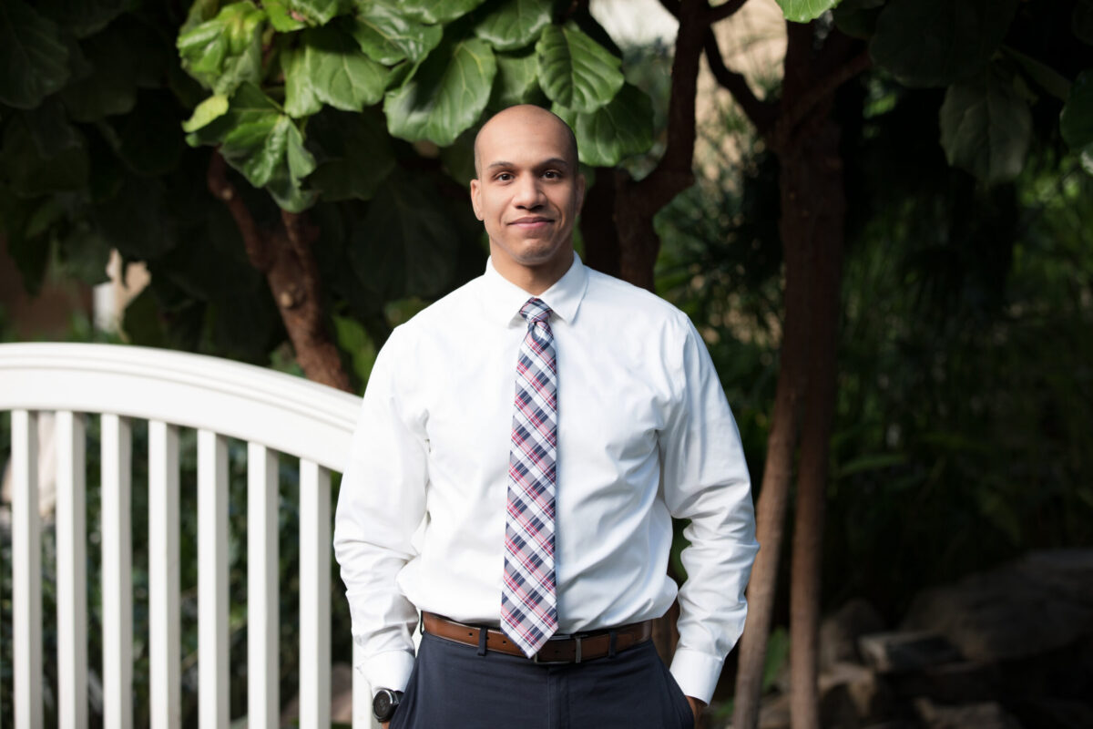 A professionally dressed man stands outdoors near a white footbridge, wearing a white dress shirt, patterned tie, and dark slacks, with greenery and trees in the background.